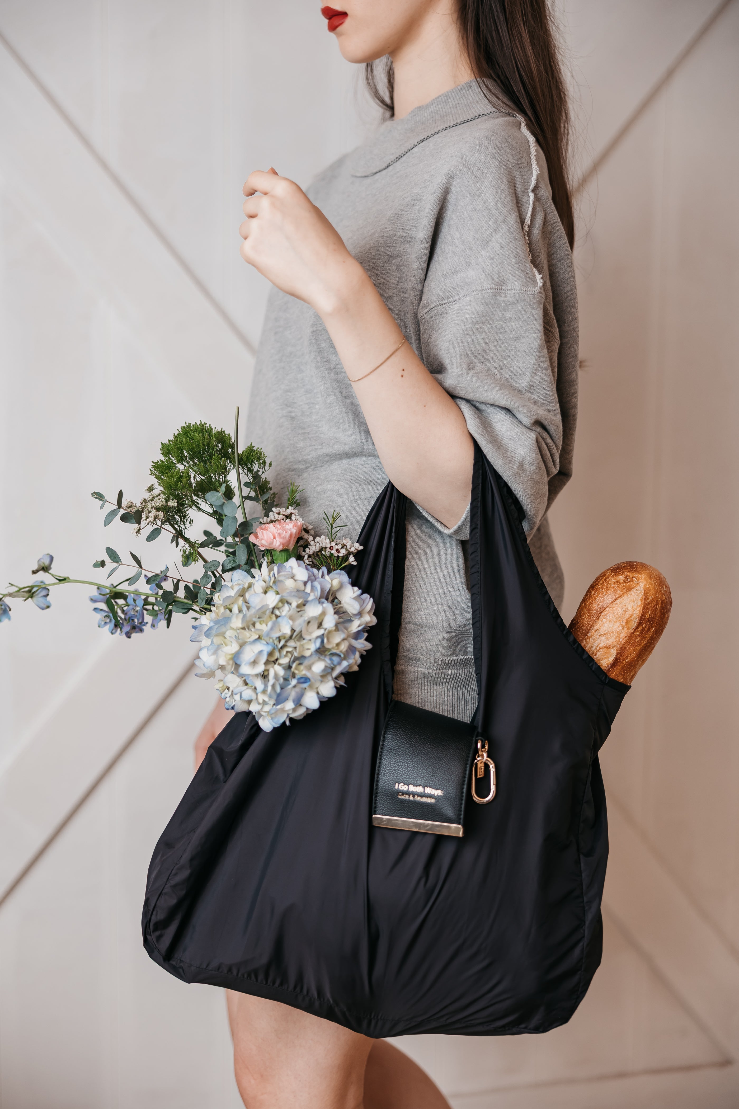 Woman holding a black reusable bag with flowers and bread against a neutral background market bag, charm, jane the brand