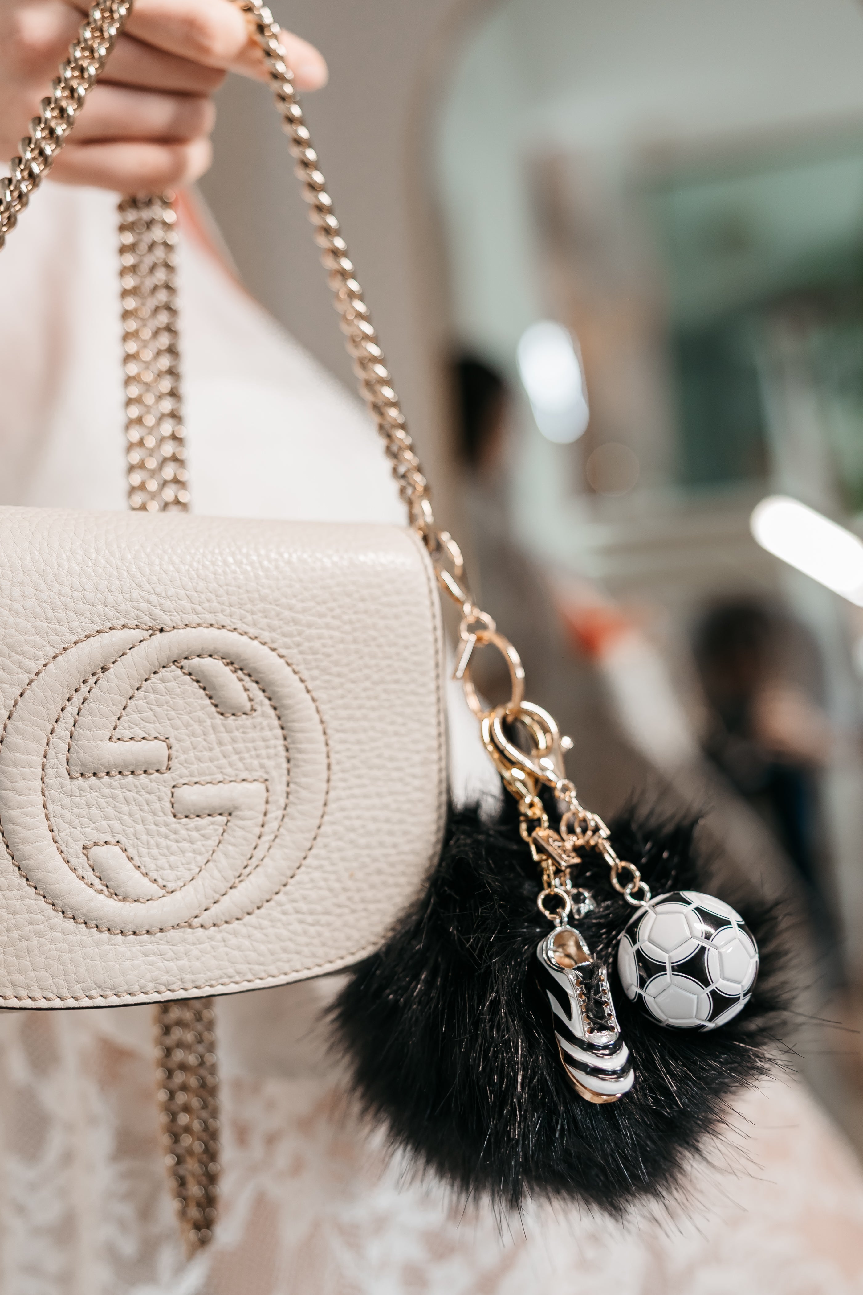 Beige handbag with decorative charms including a black fuzzy pom and a soccer ball charm.
