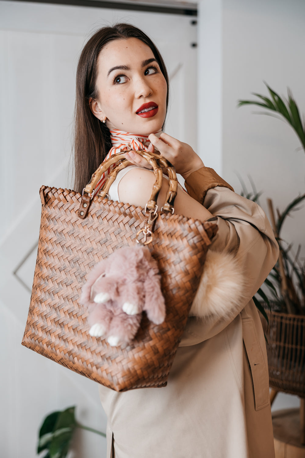 Woman holding a woven handbag with a pink plush bunny, wearing a beige coat indoors. bag charms