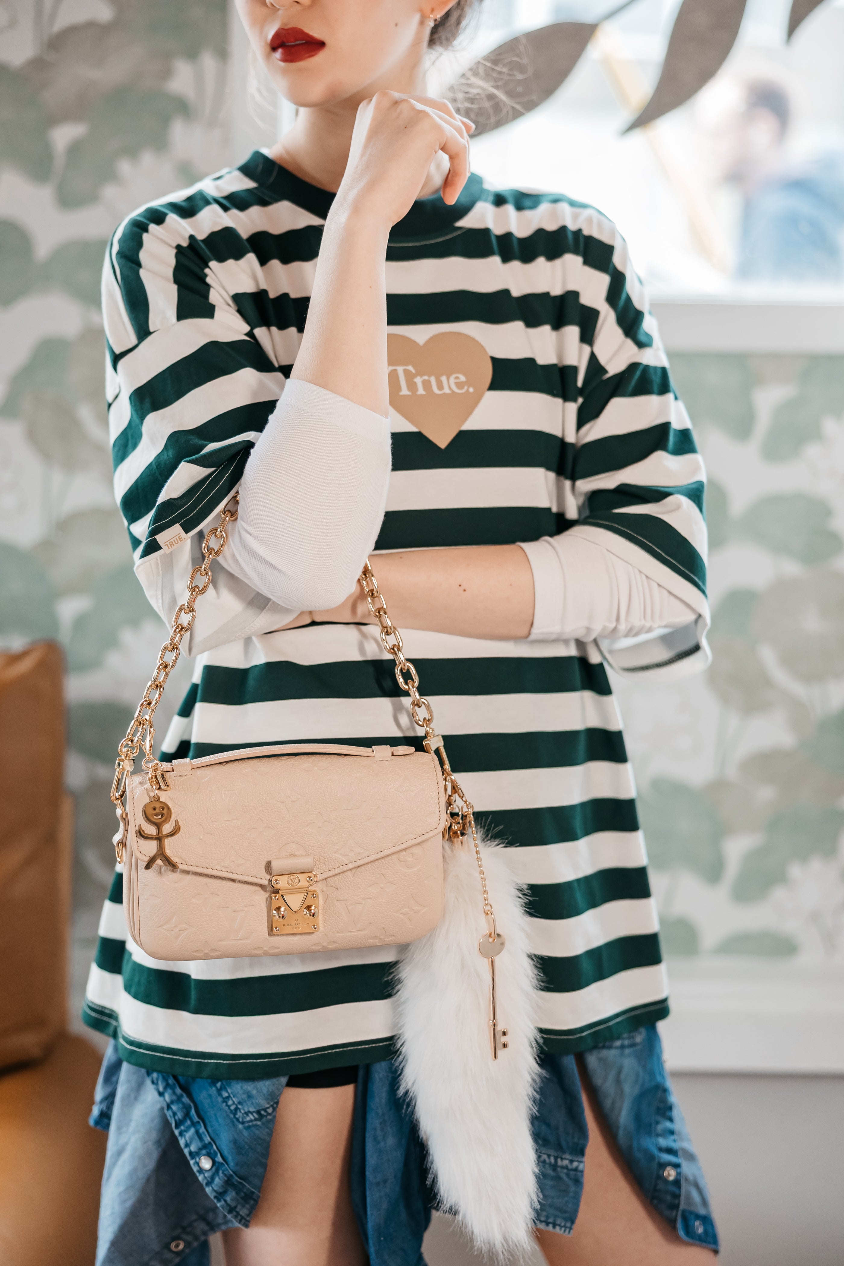 Person wearing a green and white striped sweater with a heart design, holding a beige handbag bag charm no fucks gold key white tail charm
