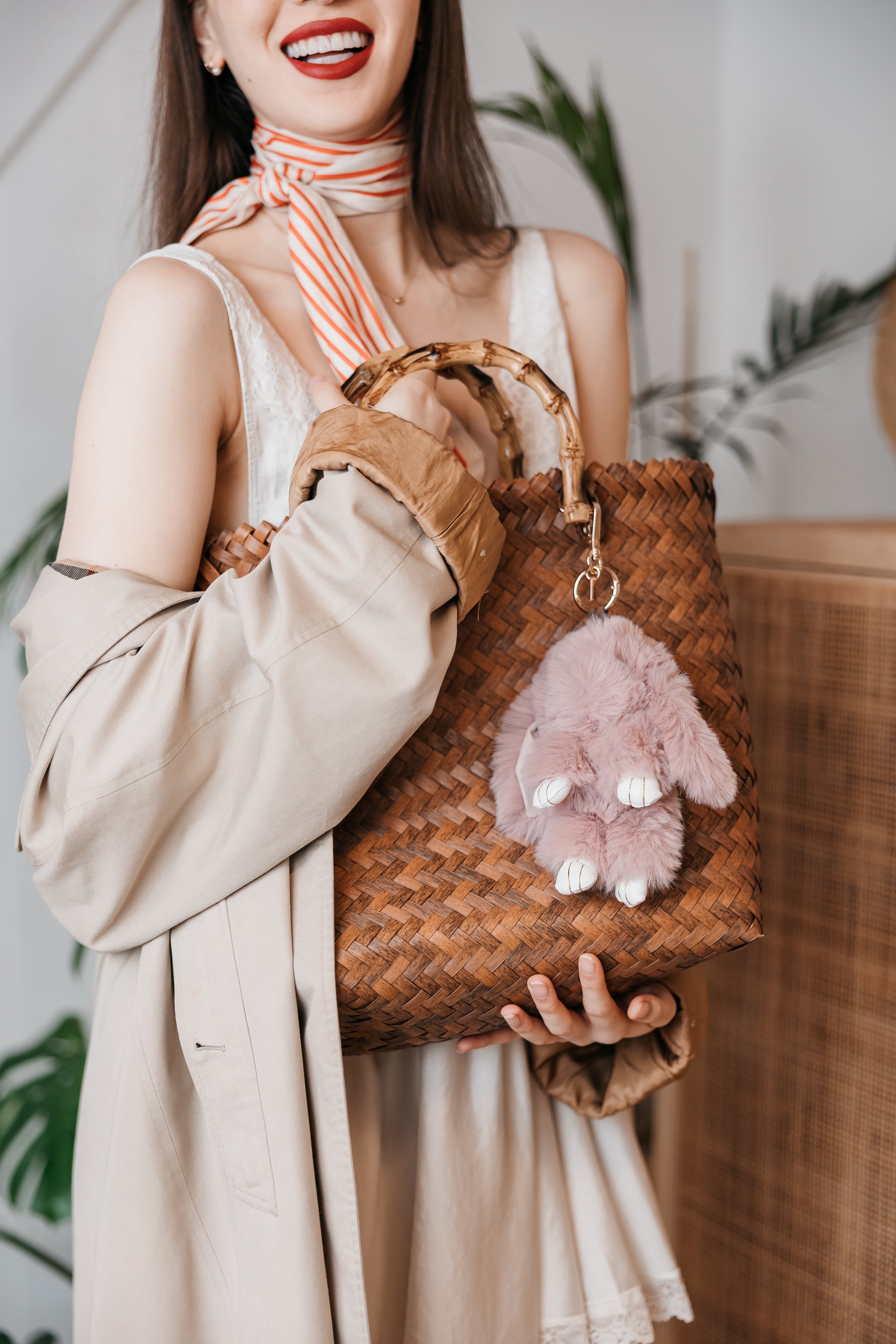Woman holding a brown woven handbag with a plush bunny charm, wearing a beige coat and striped scarf.